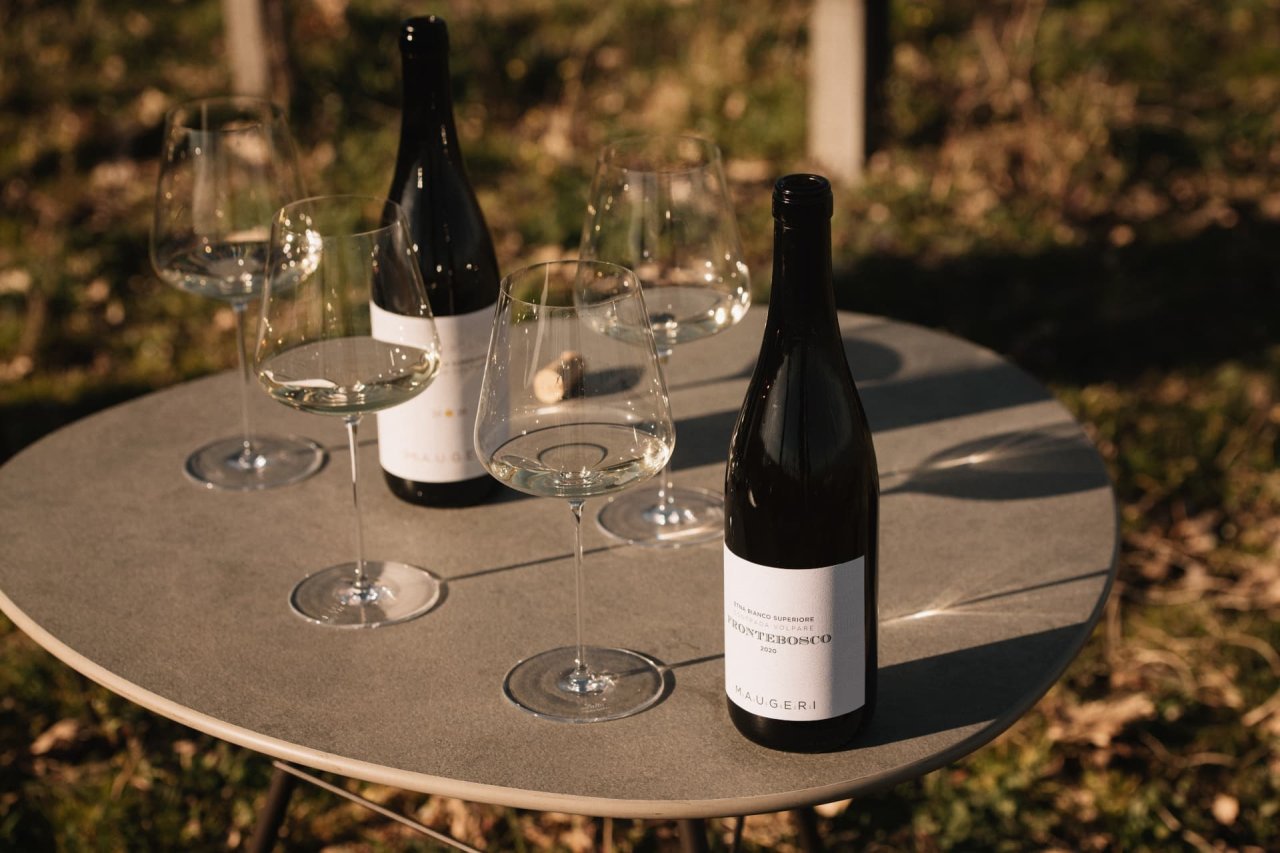Four glasses of white wine and two Maugeri bottles, one of them Frontebosco 2020, on a round table among the vines.