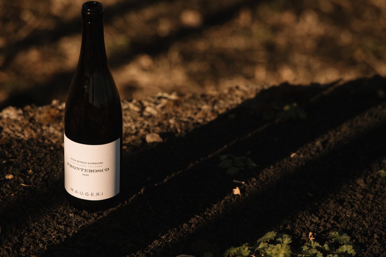 A bottle of Maugeri Frontebosco 2020 Etna Bianco Superiore standing on dark volcanic soil in low sunlight.