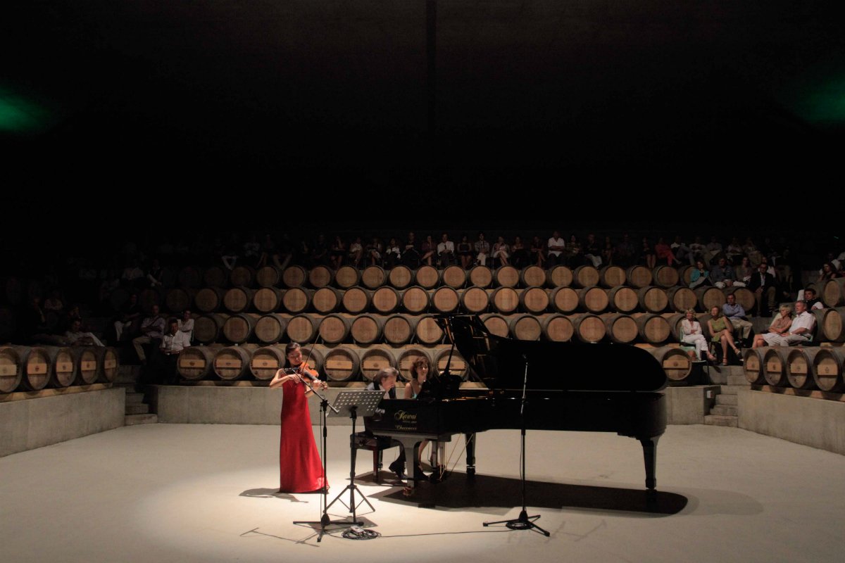 A violinist in a red dress and a pianist at a grand piano play to an audience seated among the barrels in the barrel amphitheatre at Rocca di Frassinello