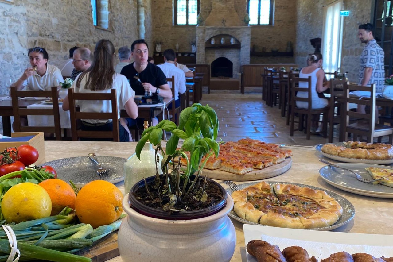 Guests at tables in the beamed dining hall of Tenute Baglio Passofondo, with pizzas and fruit in the foreground