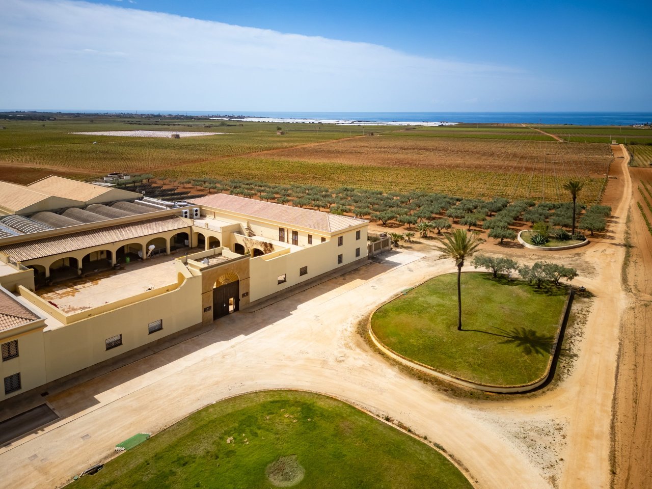 Aerial view of the cream-walled cellar buildings at Tenuta Gorghi Tondi, with olive trees, vineyards and the sea beyond.
