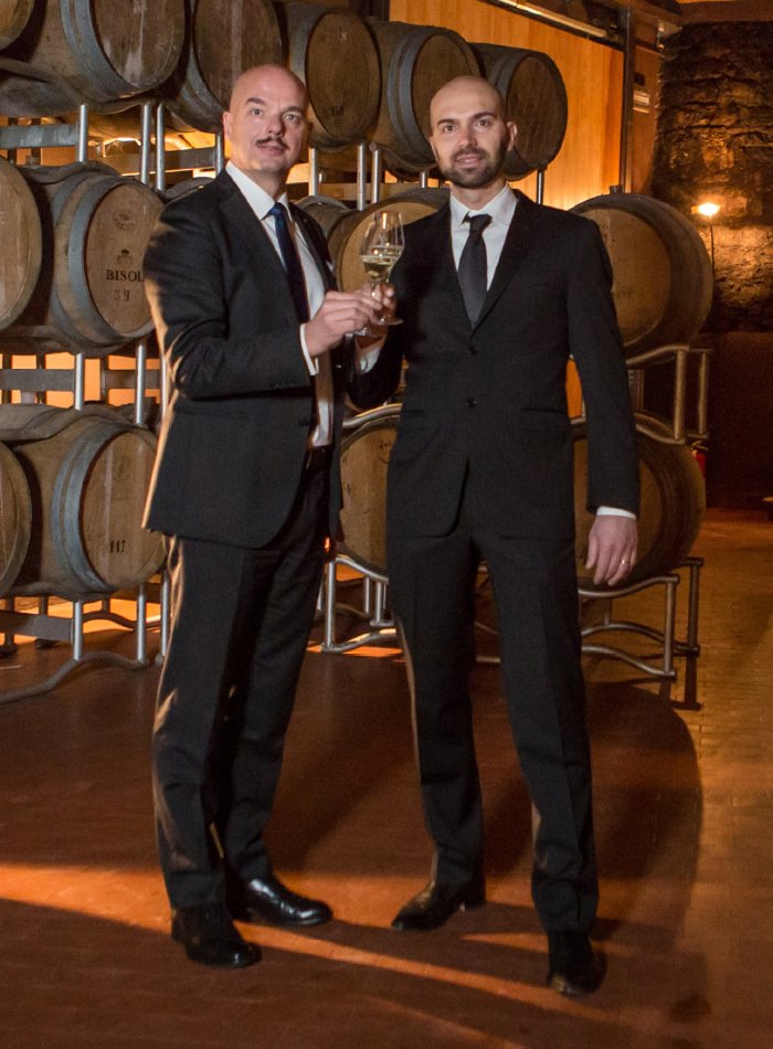 Two men of the Bisol family in dark suits holding glasses in the barrel cellar