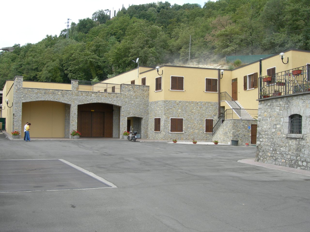 The La Carraia winery building, with yellow walls and stone arches, around a paved yard below a wooded hill