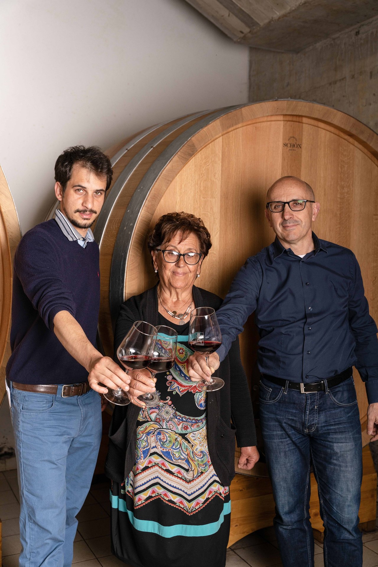 Emilio, Lidia and Stefano of Tenuta La Viola raising glasses of red wine in front of a large oak cask in the cellar