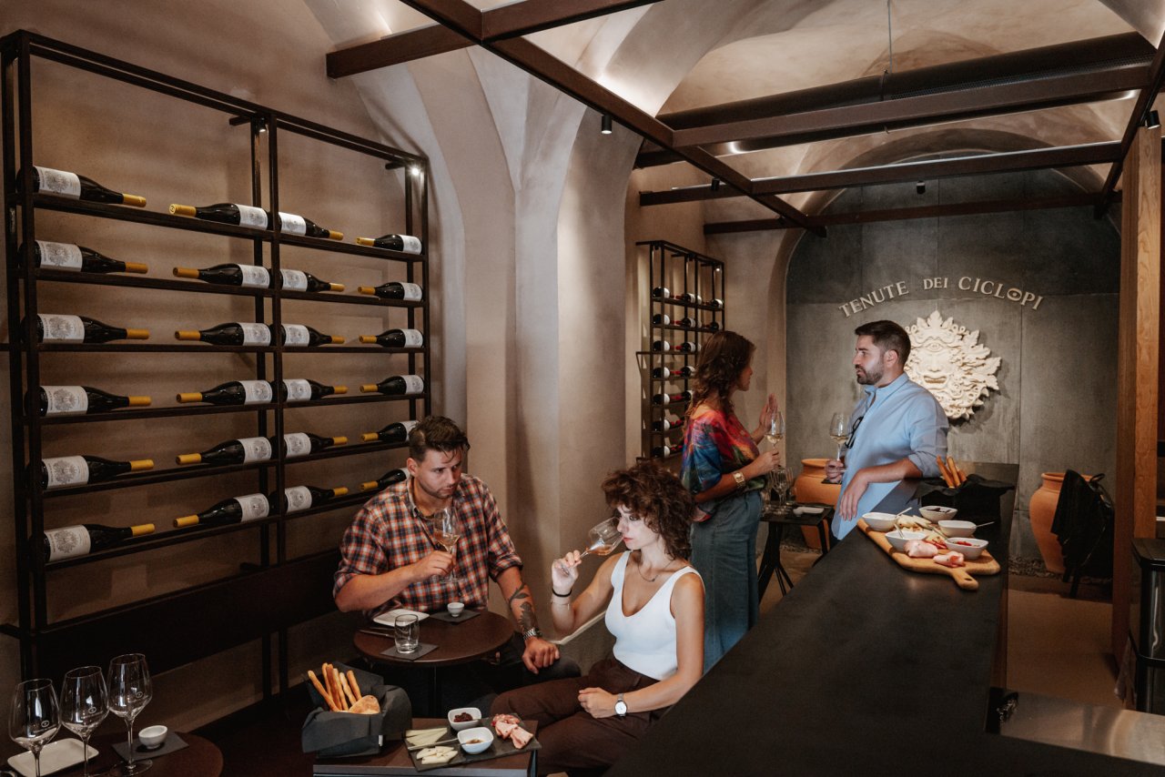 Guests at a table in the Tenute dei Ciclopi tasting room in Catania, with the bottle wall on the left and the estate's name and one-eyed relief on the back wall