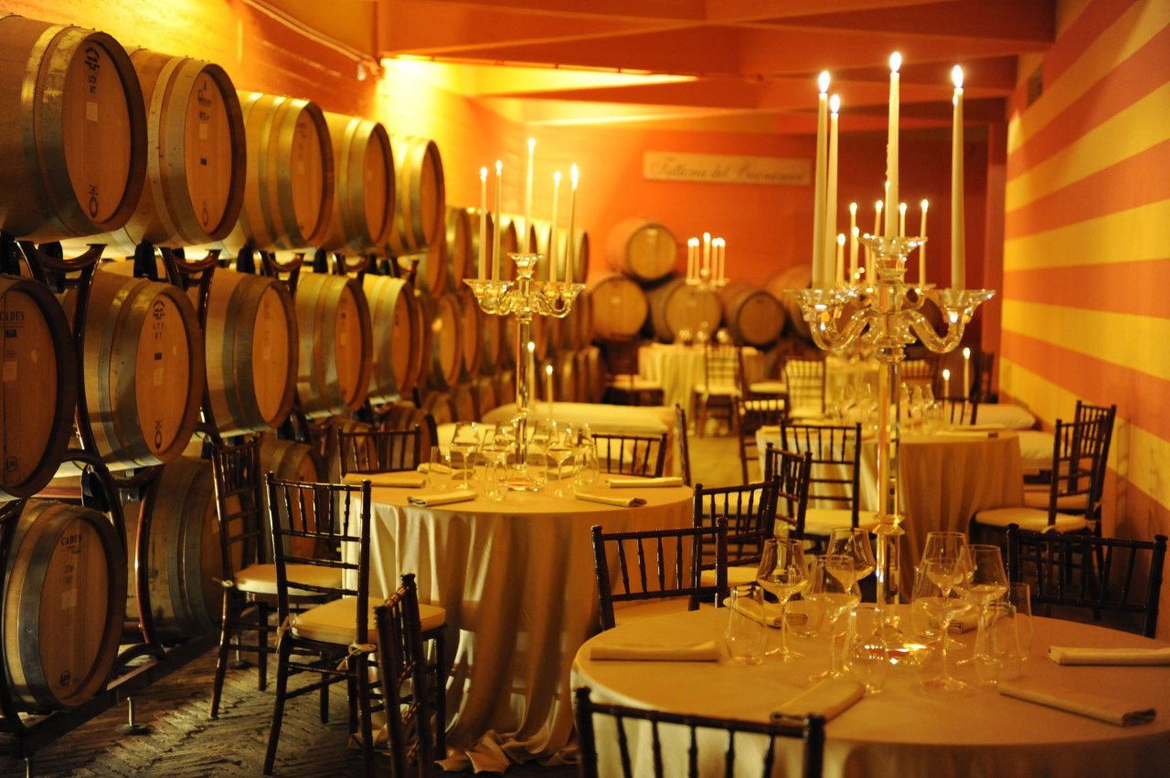 Barrel cellar at Tenuta del Buonamico laid with round dinner tables and tall candelabra, a Fattoria del Buonamico sign on the far wall