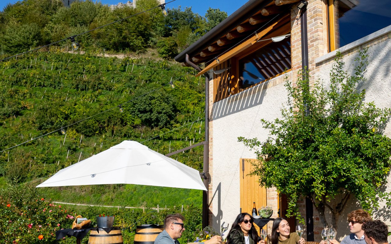Guests raising glasses under a white parasol at Follador's Rustico, with vines and a tower on the hill behind
