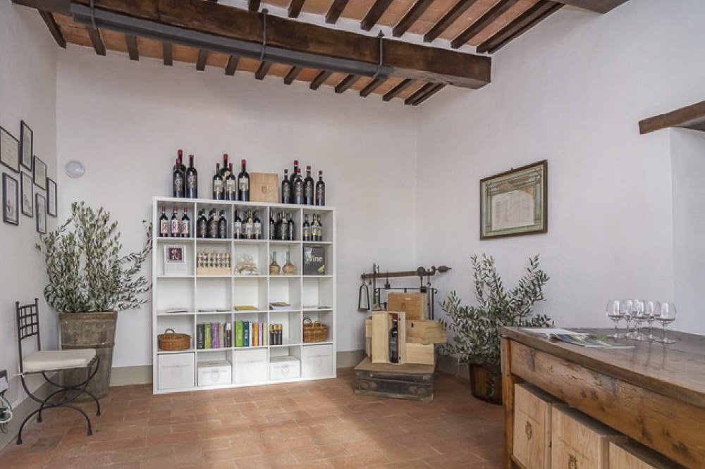 A room at Castello di Montegiove with bottles on white shelves, olive trees in pots and wine glasses on a wooden counter