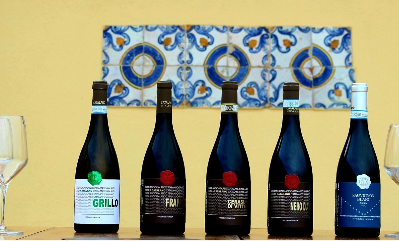 Five Catalano Viticoltori bottles in a row on a table against painted Sicilian tiles: Grillo, Frappato, Cerasuolo di Vittoria, Nero d'Avola and Sauvignon Blanc