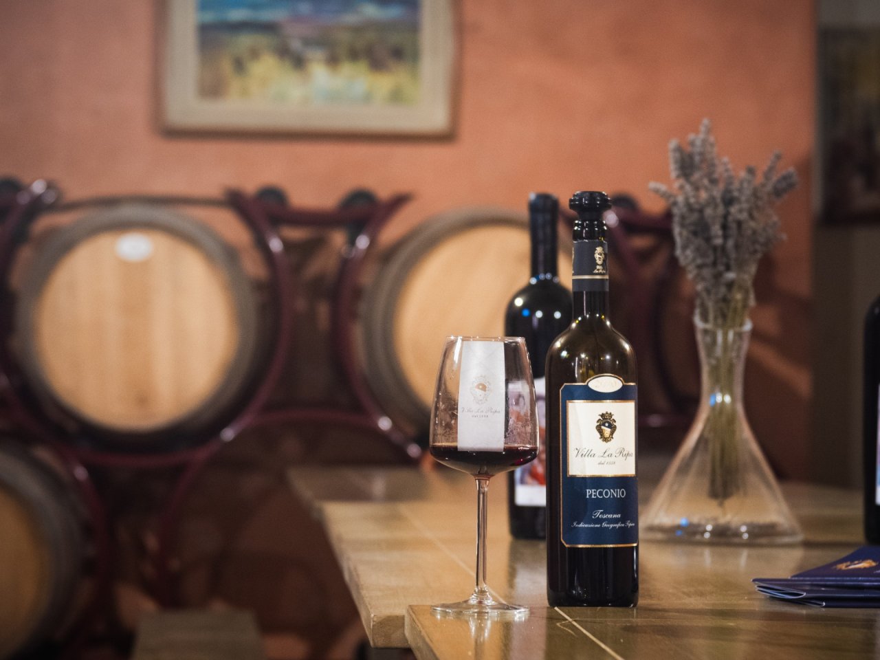 A bottle of Villa La Ripa Peconio beside a glass of red wine, with barrels behind