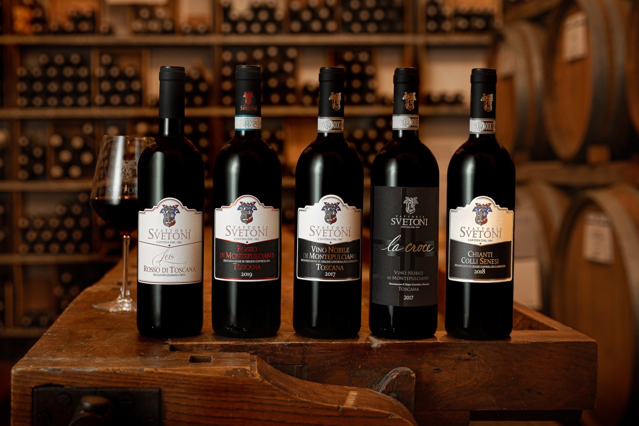 Five Fattoria Svetoni wines lined up on a wooden stand in the cellar, with a glass of red beside them
