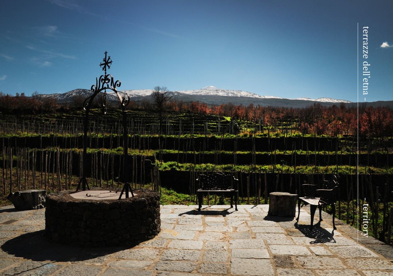 Stone terrace with an old well and iron benches at Terrazze dell'Etna, vineyards and snow-capped Etna beyond