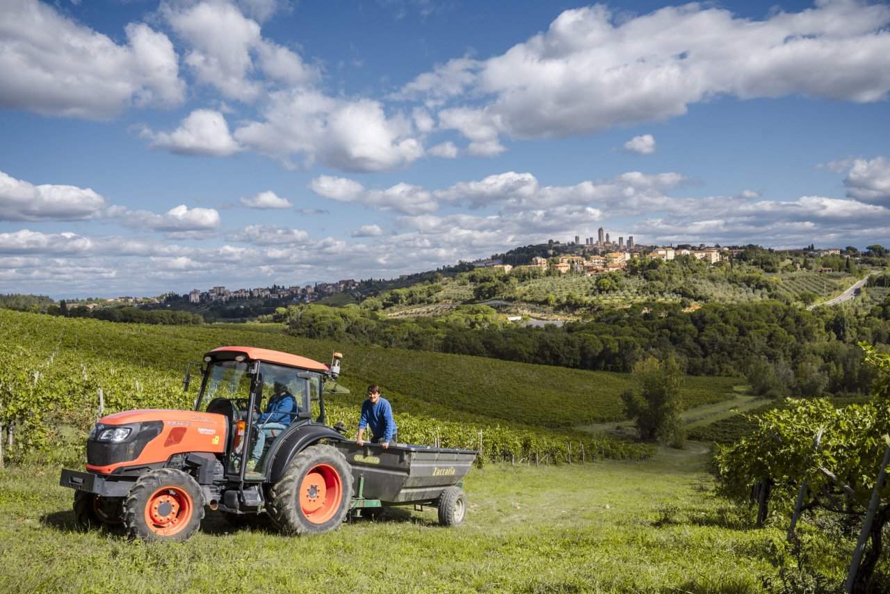 An orange tractor and trailer in a Palagetto vineyard, with the towers of San Gimignano on the hill behind