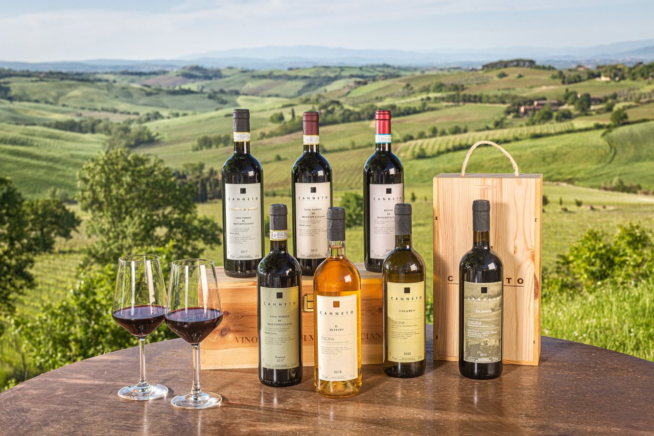 Canneto wines lined up on a table outdoors, with two glasses of red and green Tuscan hills behind